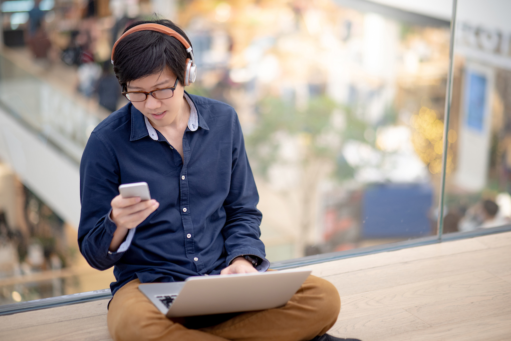 Young male student wearing headphones and looking at phone while working on laptop balanced on his crossed legs.