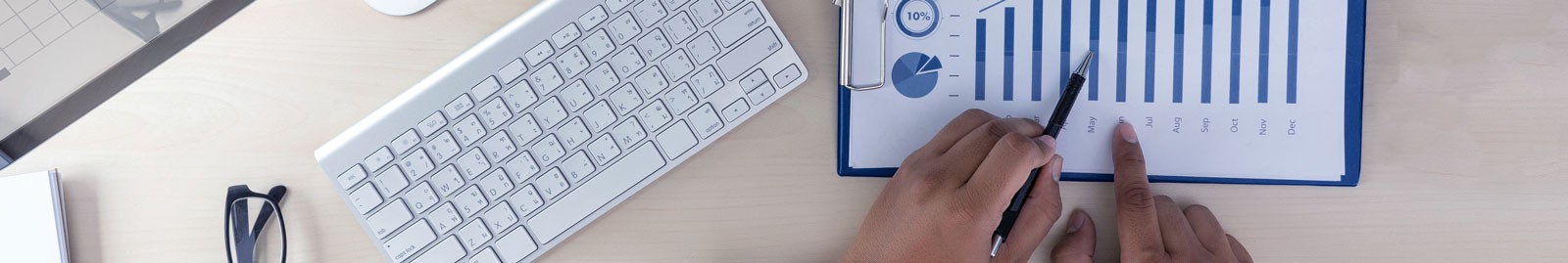 Hands look over a chart with a pen, while glasses and a keyboard sit on the desk.