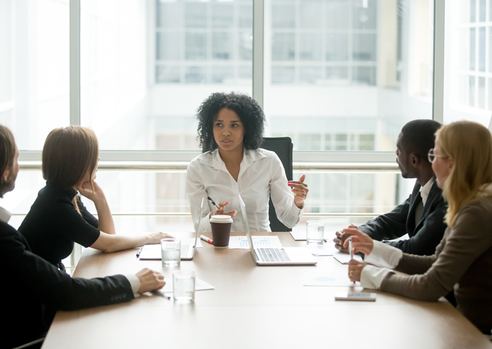 A black woman sits at the head of a conference table, leading a meeting with colleagues.