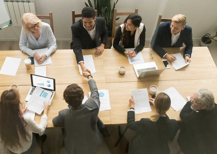 Viewed from above, eight people sit at a table. two are shaking hands over the table.