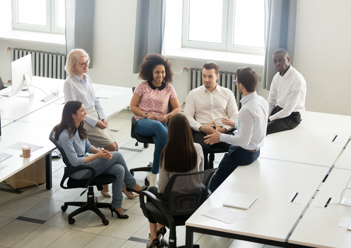 A group of seven people talk at their desks.