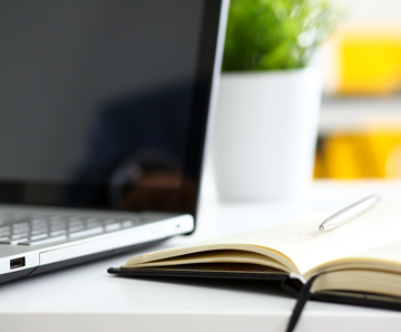 A journal lies open on a desk next to a silver laptop. A pen is on top of the journal.